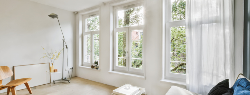 a living room with white walls and wood flooring, two large windows in the wall is covered with sheer curtains