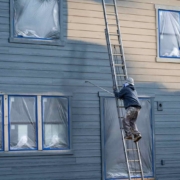 Professional painter in hard hat and air respirator on extension ladder with paint sprayer painting back exterior of house, job site for new housing development