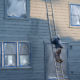 Professional painter in hard hat and air respirator on extension ladder with paint sprayer painting back exterior of house, job site for new housing development