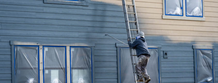 Professional painter in hard hat and air respirator on extension ladder with paint sprayer painting back exterior of house, job site for new housing development