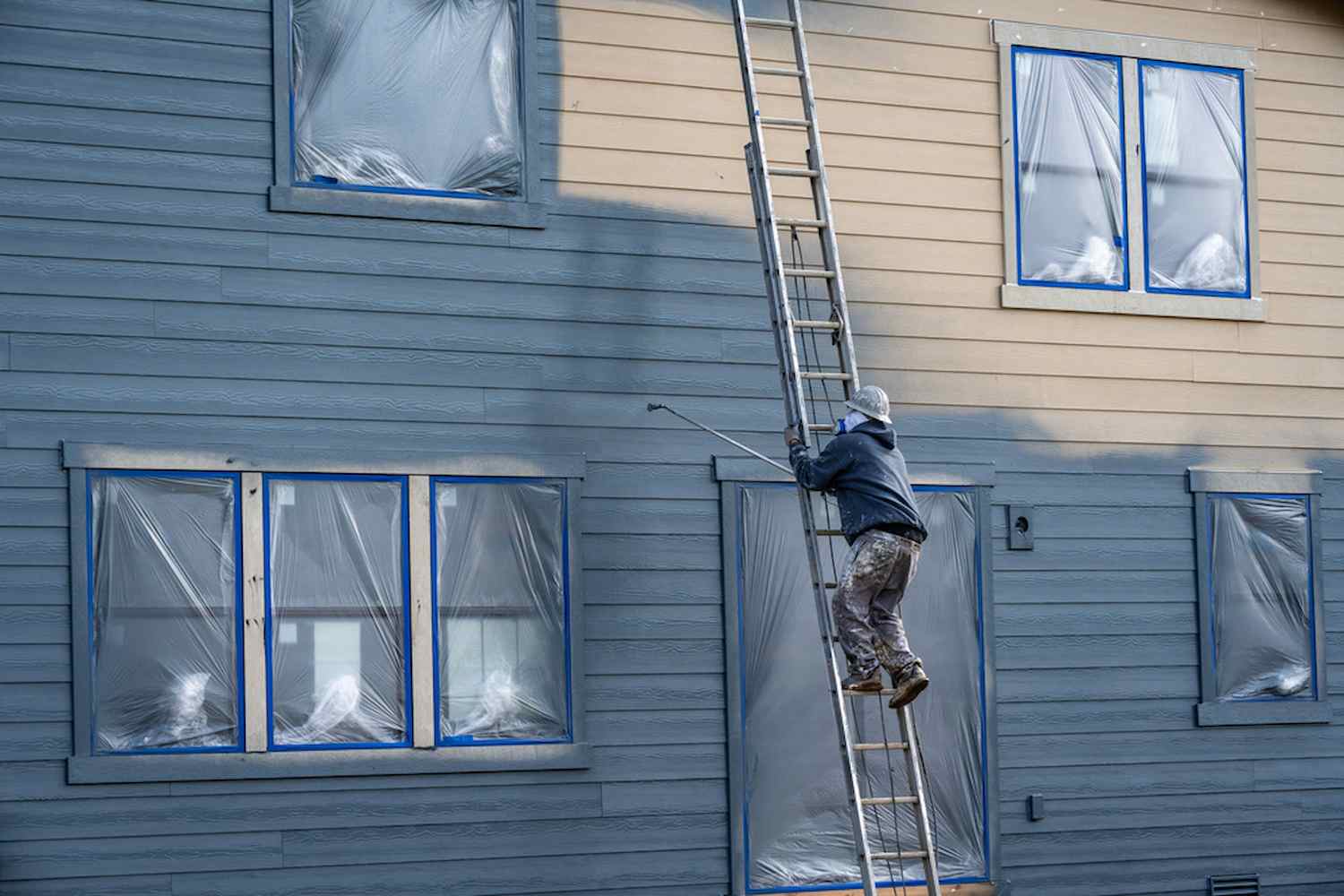 Professional painter in hard hat and air respirator on extension ladder with paint sprayer painting back exterior of house, job site for new housing development