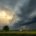The image depicts a dramatic scene of storm clouds looming over a Missouri landscape, with dark, swirling formations hinting at impending rain. In the foreground, the lush greenery of fields contrasts with the ominous sky, creating a striking visual of nature's power.